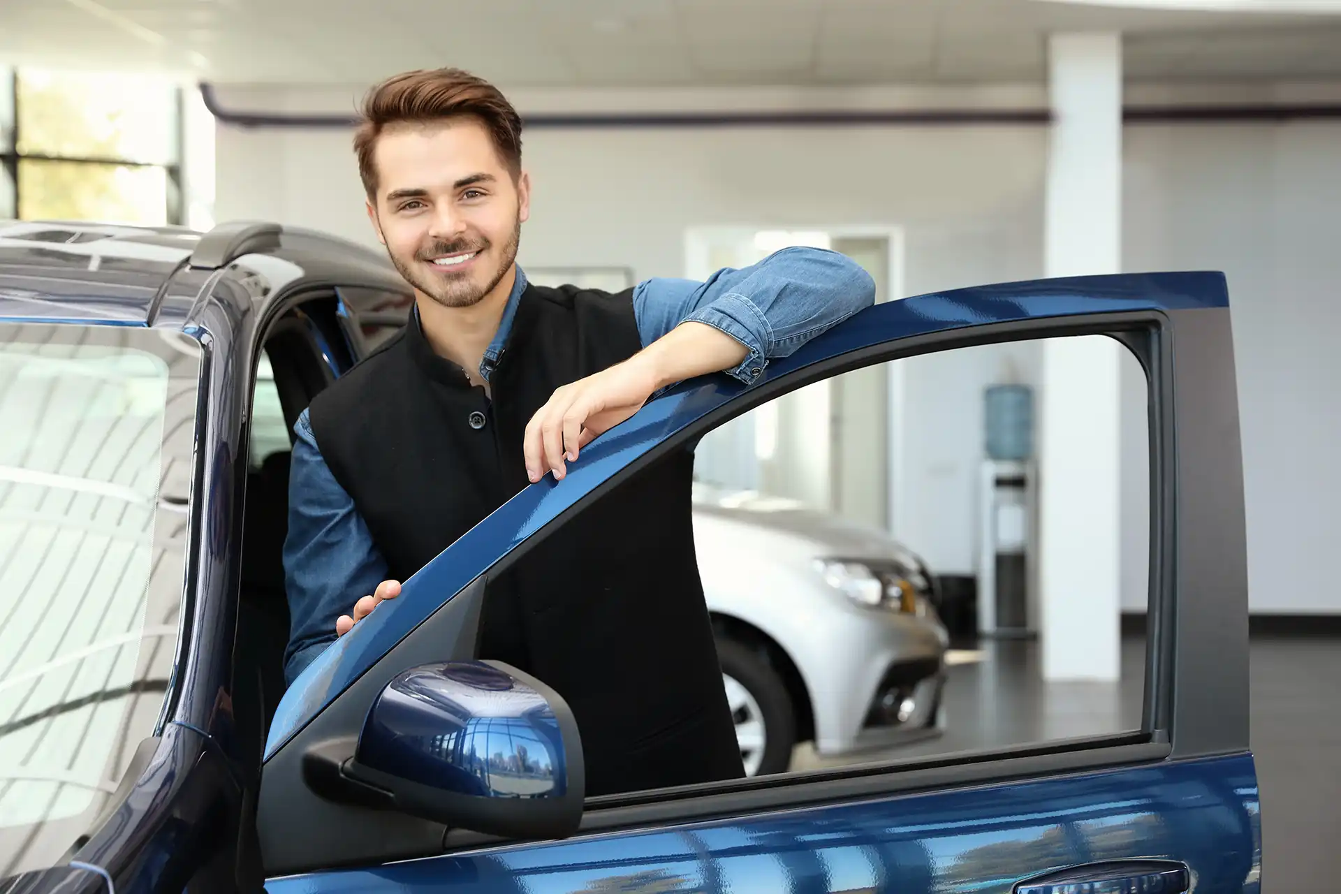 junger Mitarbeiter im Autohaus lehnt an geöffneter Autotür und lächelt in die Kamera - Stellenangebot Kfz-Serviceberater / Automobil-Serviceberater